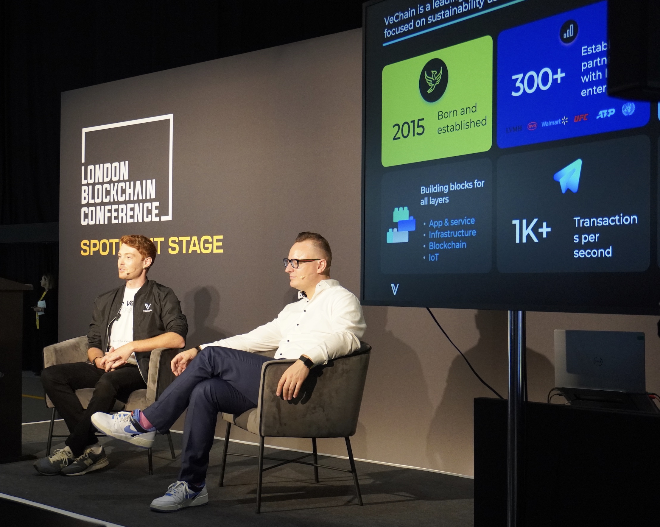 Jake Campton speaking on the London Blockchain Conference Spotlight Stage in 2024, with Bernhard Kronfellner (Partner, Boston Consulting Group) listening. The VeChain pitch deck visible on screen behind them.